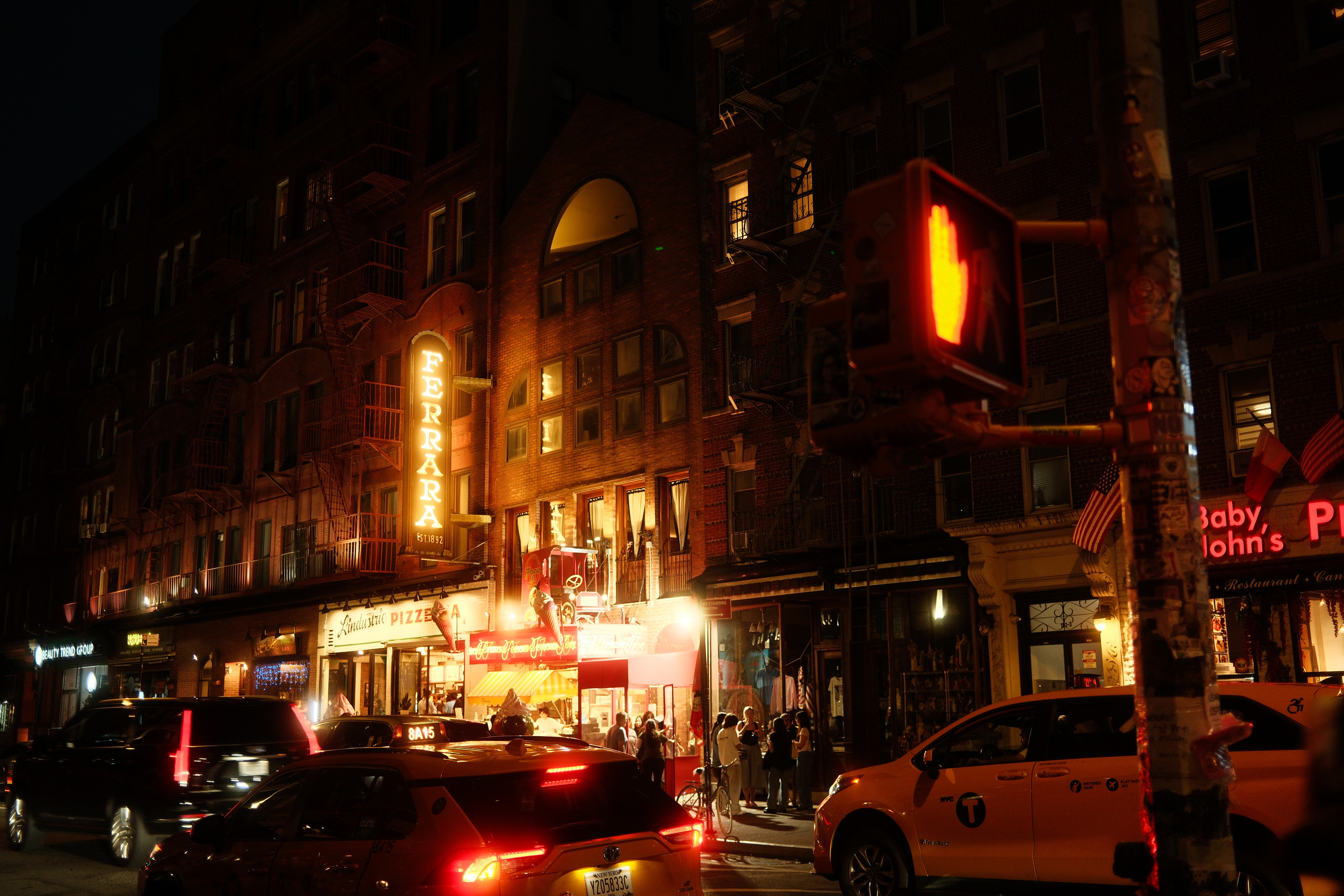 Neon signage above a Little Italy street corner in New York at night, taxis passing in the foreground.