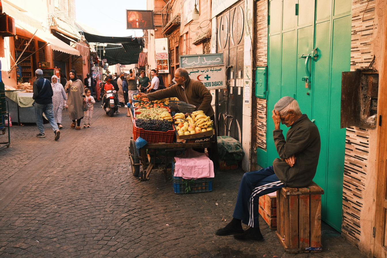 A fruit cart in a Marrakech medina lane at golden hour, an older man resting on a crate beside a green door.