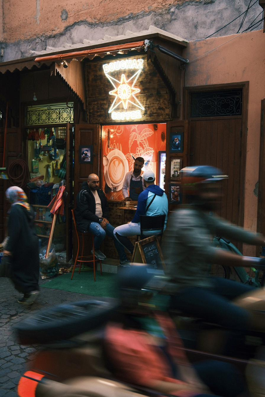 A neon-lit coffee window in the medina, two customers seated as a scooter blurs past.