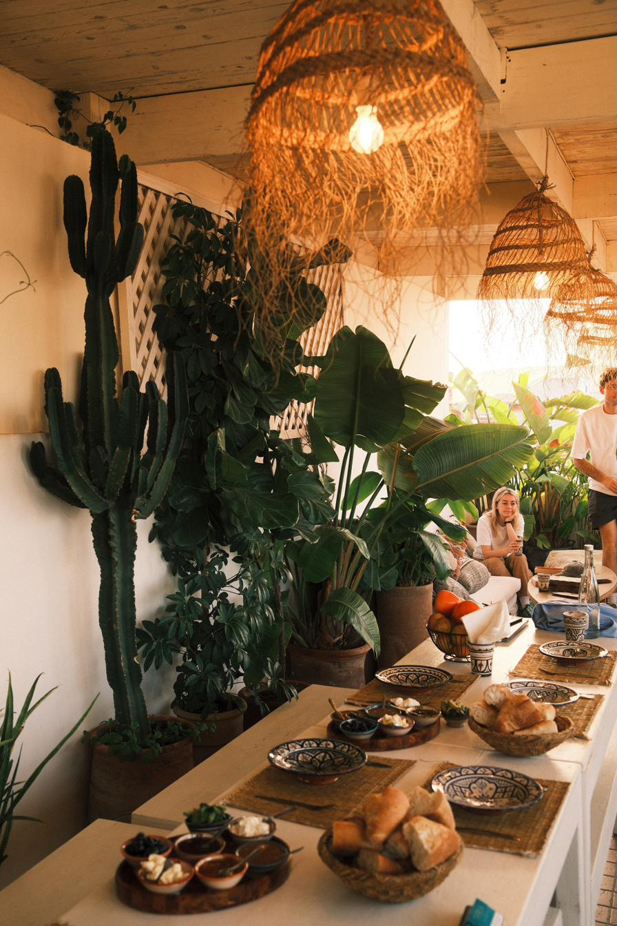 A long breakfast table on a plant-filled rooftop, woven lamps overhead and guests in the late sun.
