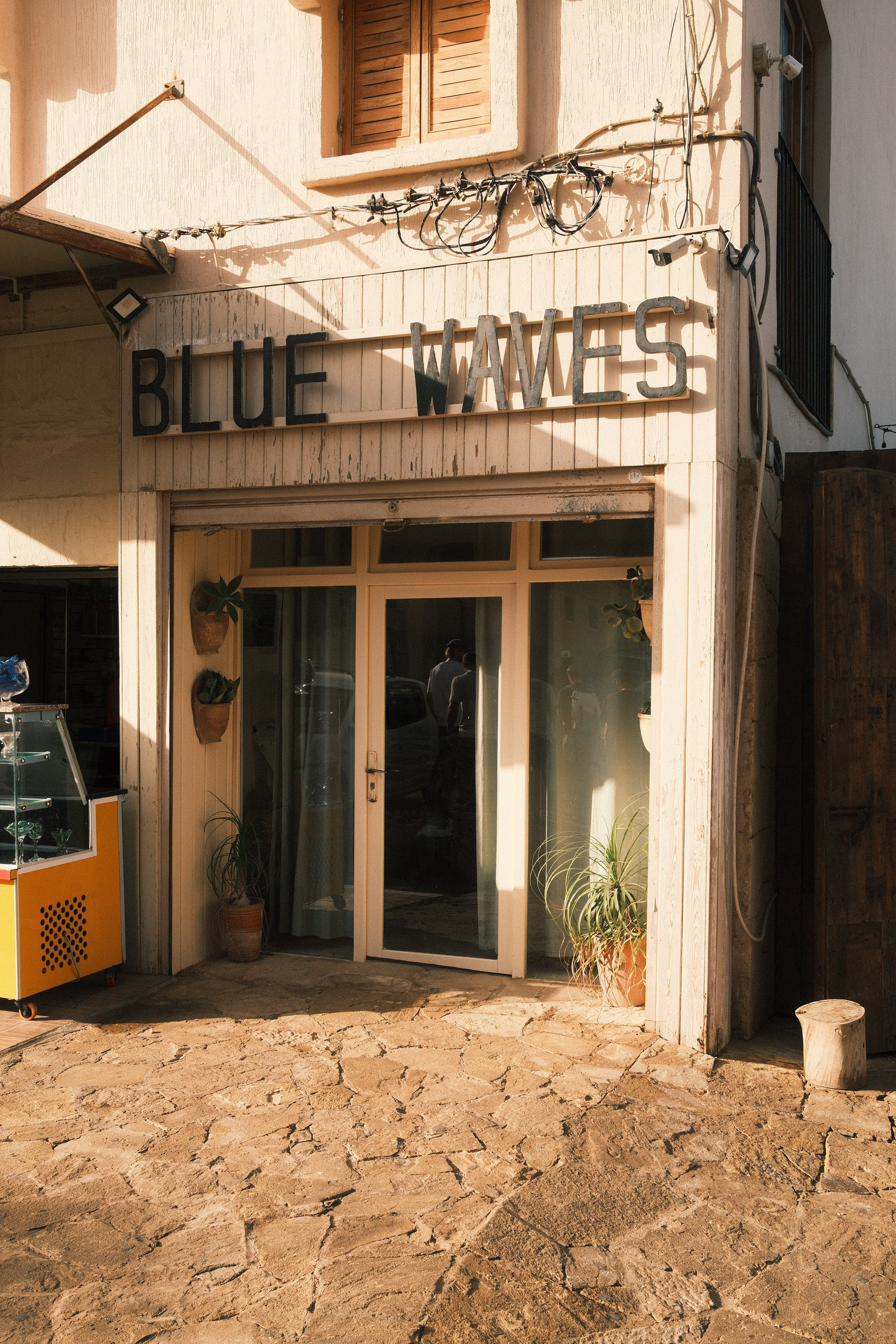 A weathered shopfront lettered Blue Waves, low sun raking across the stone forecourt.