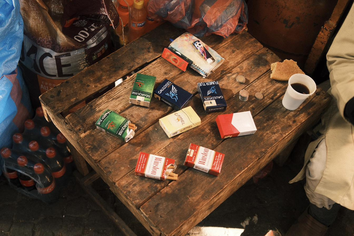 Cigarette packs, coins and a cup of coffee laid out on a wooden kiosk counter.