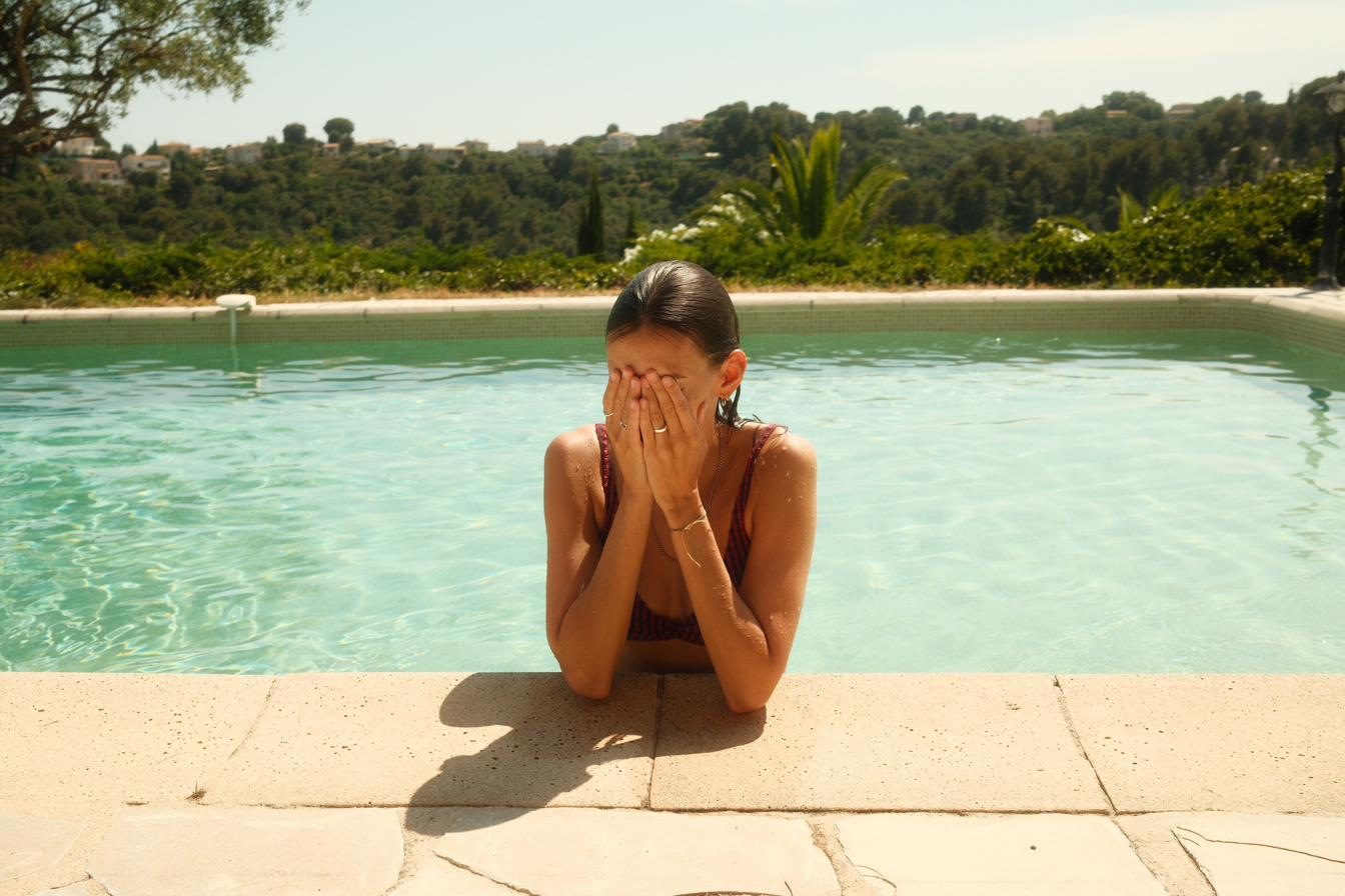 A woman at the edge of a hillside pool, hands over her face, hills and cypresses behind.