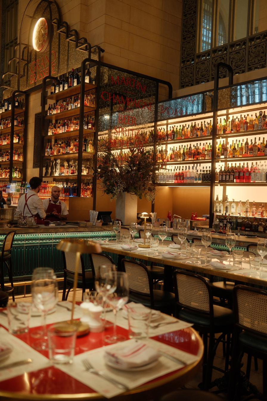 A brasserie bar under a vaulted hall, bottles backlit along the shelves, two bartenders talking.