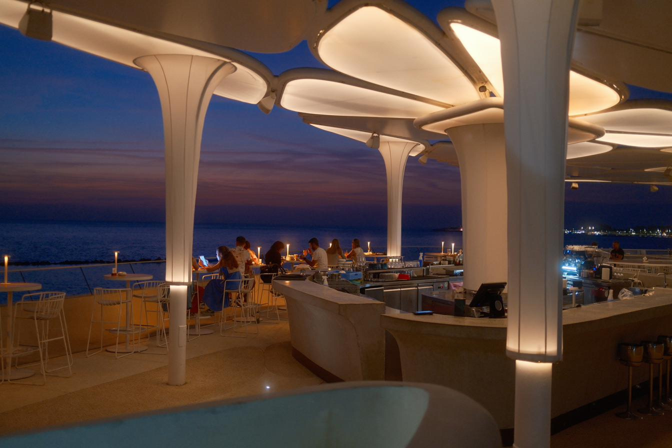 A seafront bar under sculpted white canopies at blue hour, candles lit along the counter.