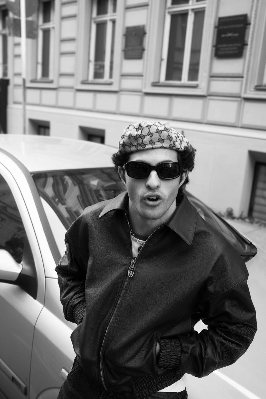 Black and white portrait of a man in a leather jacket and headscarf leaning against a car.