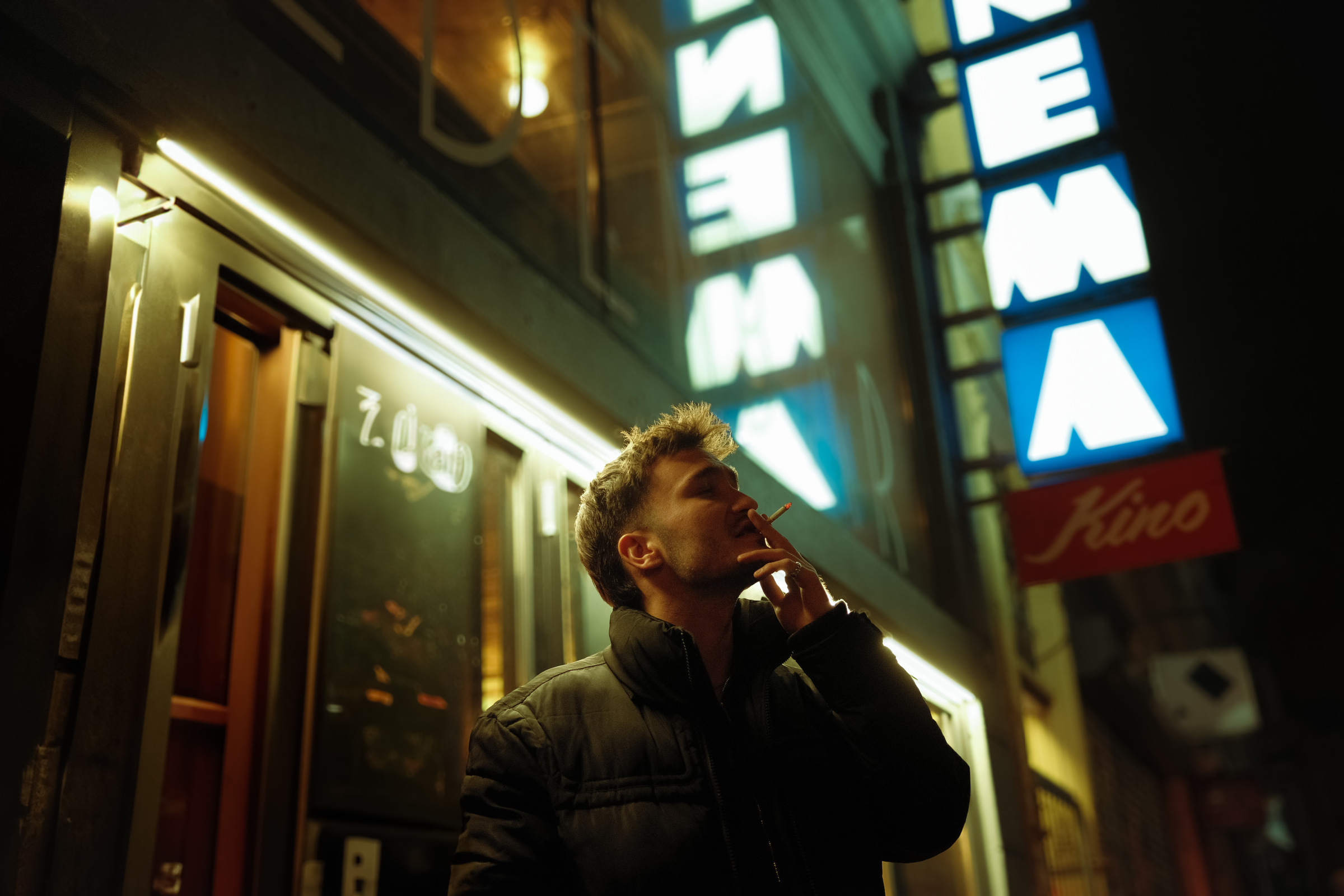 A man smoking outside a cinema at night, lit from the side, the blue neon letters of the marquee glowing above him.