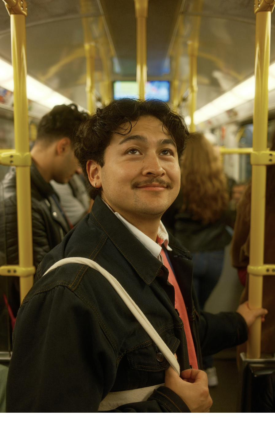 A young man with a moustache holding a pole on a crowded underground carriage, looking up.