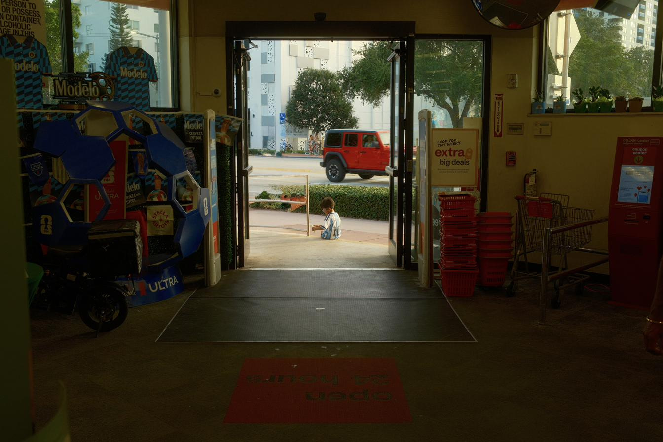 Seen from inside a drugstore, a boy in a football shirt sits on the kerb in the doorway light.