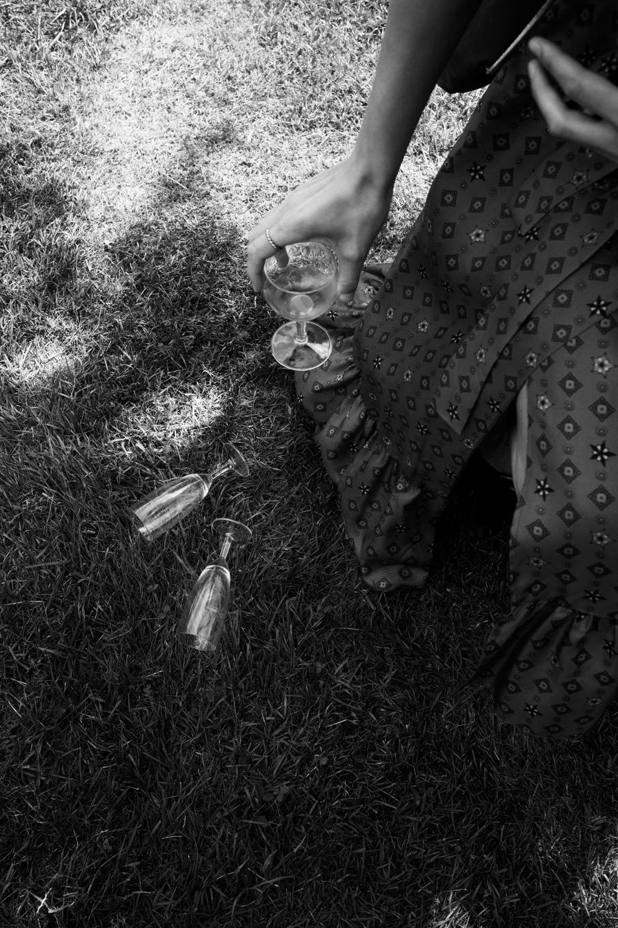 Black and white overhead frame of a woman in a printed dress collecting wine glasses from the grass.