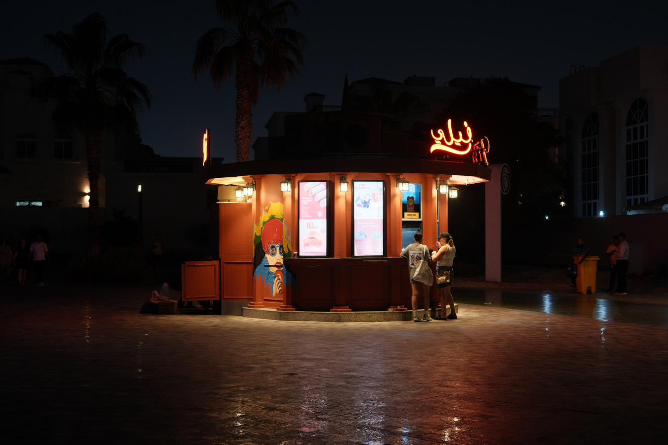 A lit kiosk under palms at night, two customers ordering, wet paving reflecting the neon.