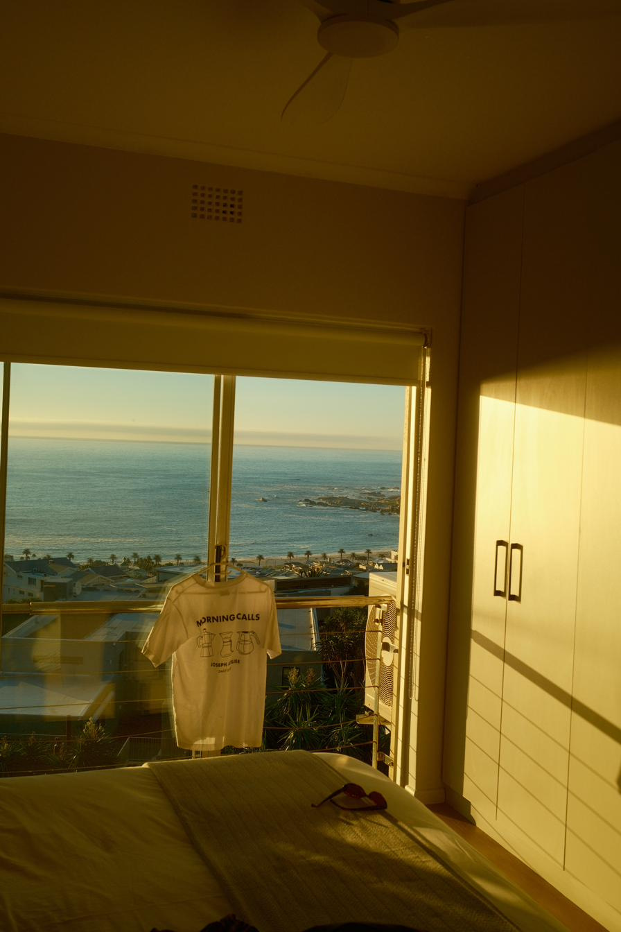 A white t-shirt hung in an open bedroom window above the sea, warm morning light on the wall.