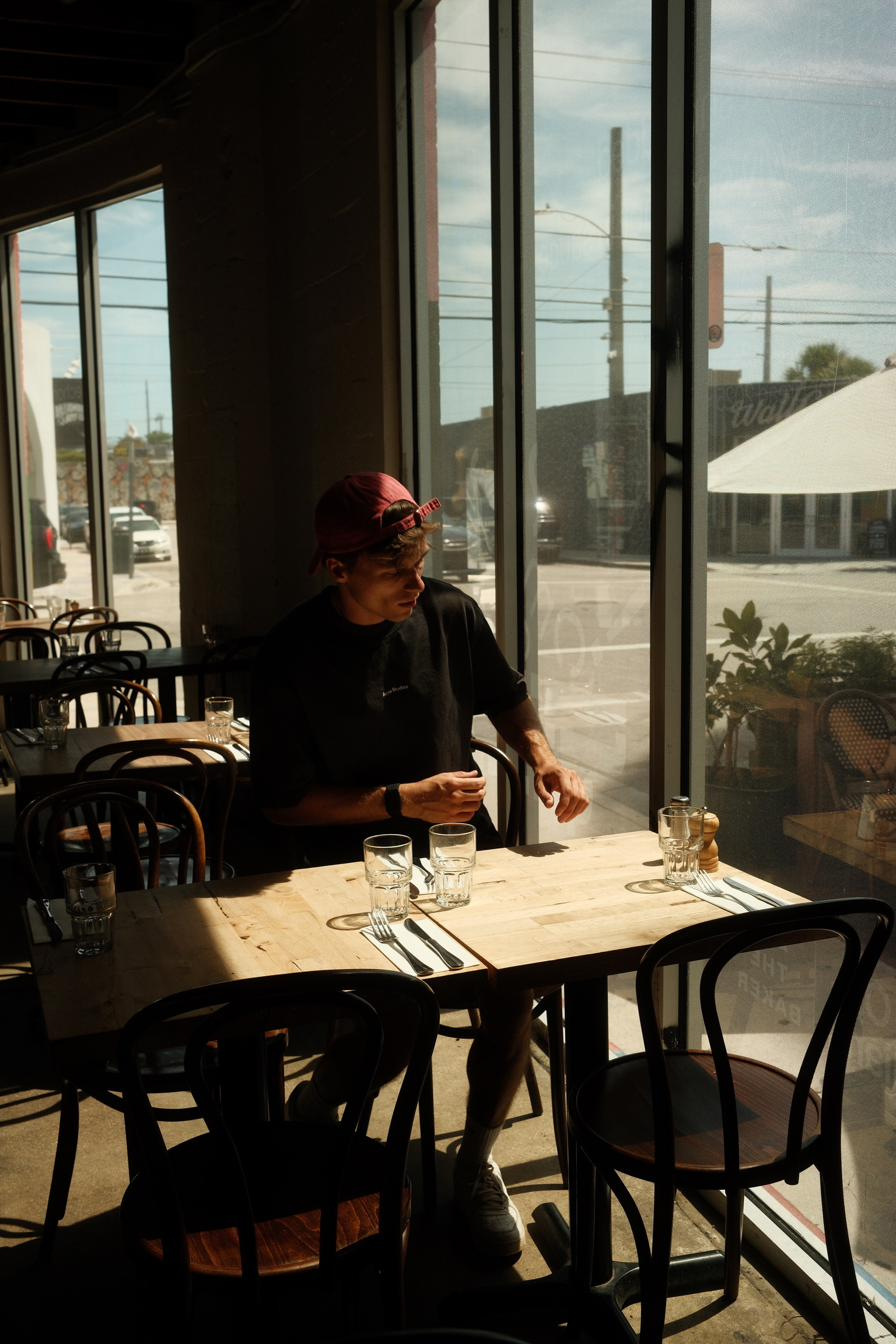 A man in a backwards cap at a window table in an empty café, hard midday light outside.